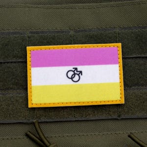 Op de afbeelding: Een rechthoekige patch met een gele rand, met een horizontaal driekleurig ontwerp van roze, wit en geel. De patch bevat een zwart symbool van twee in elkaar grijpende mannelijke symbolen. De patch is bevestigd aan een groene stof.