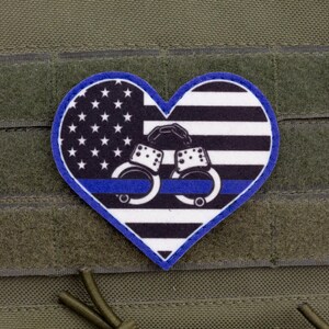 May include: A heart-shaped patch featuring a stylized American flag design with black and white stripes, a blue field with white stars, and a thin horizontal blue line across the center. Centered on the patch are white handcuffs.
