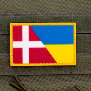 Könnte beinhalten: Ein rechteckiger Aufnäher mit den Flaggen Dänemarks und der Ukraine. Die dänische Flagge mit weißem Kreuz auf rotem Grund befindet sich links. Die ukrainische Flagge mit blauen und gelben Streifen befindet sich rechts. Der Aufnäher hat einen gelben Rand.