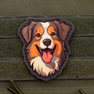 Könnte beinhalten: Ein farbenfroher Aufnäher mit einem lächelnden Hund mit braunem, orangefarbenem und weißem Fell. Die Zunge des Hundes ist sichtbar, und der Aufnäher befindet sich auf einem grünen Hintergrund mit Klettverschluss.