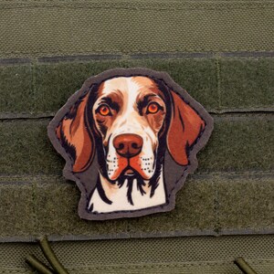 Op de afbeelding: Een patch met een gedetailleerde illustratie van een hondenhoofd, met bruine en witte vacht en oranje ogen. De patch is geplaatst tegen een groene achtergrond met een klittenbandachtige textuur.