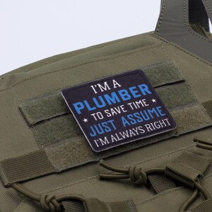 K&ouml;nnte beinhalten: Olivgr&uuml;ne taktische Weste mit einem rechteckigen Aufn&auml;her. Der Aufn&auml;her tr&auml;gt die Aufschrift "I'M A PLUMBER TO SAVE TIME JUST ASSUME I'M ALWAYS RIGHT" in wei&szlig;er und blauer Schrift. Die Weste hat mehrere Riemen und einen Klettverschluss.