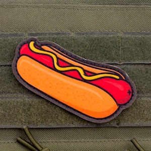 May include: A colourful patch featuring a cartoon hot dog with a bright orange bun, red sausage, and yellow mustard. The patch is set against a green background with a hook and loop fastener.