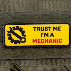 Könnte beinhalten: Ein gelbes rechteckiges Abzeichen mit dem Text "TRUST ME I'M A MECHANIC" in Schwarz und Rot. Es zeigt ein schwarzes Schraubenschlüssel- und Zahnradsymbol. Das Abzeichen ist an einem grünen Stoff befestigt.