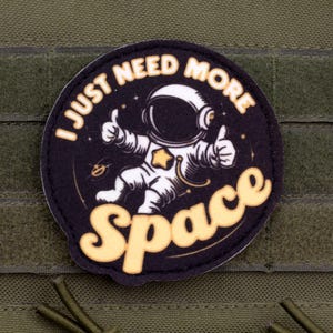 May include: A round patch featuring an astronaut giving a thumbs-up, with the text "I JUST NEED MORE SPACE" in white and yellow. The patch is on a green fabric background.