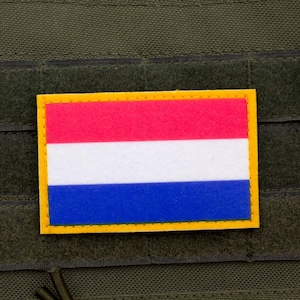 May include: A rectangular patch displaying the Dutch flag, with red, white, and blue horizontal stripes, edged in yellow. The patch is attached to a dark green fabric, possibly part of a tactical vest or similar equipment.