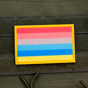 May include: A rectangular patch with a yellow border and a horizontal striped design in red, pink, light gray, blue, and yellow. The patch is affixed to a dark green fabric background with velcro-like strips.