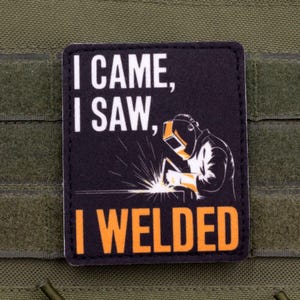 Pode incluir: Patch preto com o texto "I CAME, I SAW, I WELDED" em branco e laranja. O patch apresenta uma ilustração de um soldador com faíscas. O patch está preso a um tecido verde.