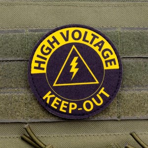 Può includere: Una toppa rotonda con le parole "HIGH VOLTAGE KEEP-OUT" in lettere gialle su sfondo blu navy. Un fulmine giallo è all'interno di un triangolo giallo. La toppa è attaccata a un tessuto verde.