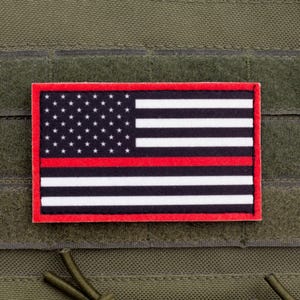 Peut inclure: Un écusson rectangulaire présentant un motif de drapeau américain stylisé avec un fond noir, des étoiles blanches, des rayures blanches et une seule rayure horizontale rouge au centre, le tout bordé d'un fin cadre rouge, fixé sur un tissu vert olive.