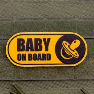 Pode incluir: Um patch amarelo e preto com o texto "BABY ON BOARD" e um gráfico de chupeta. O patch é retangular com cantos arredondados e está preso a um fundo de tecido verde.