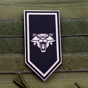 May include: A white and black patch with a white silhouette of a snarling cat's head on a black background. The patch is shaped like a shield with a white border.