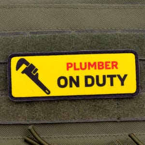 Può includere: Una toppa rettangolare con sfondo giallo e bordo nero. La toppa presenta una grafica di chiave inglese nera e le parole "PLUMBER ON DUTY" in rosso e nero. La toppa è attaccata a un tessuto verde.