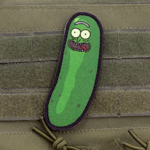 Puede incluir: Un parche verde de estilo caricatura con forma de pepinillo con una expresión de sorpresa. El parche tiene un borde negro y está adherido a un chaleco táctico con paneles de velcro. El pepinillo tiene ojos grandes y la boca abierta.