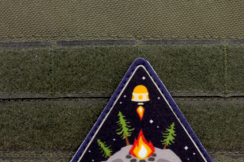 Może przedstawiać: Tr&oacute;jkątna naszywka przedstawiająca nocną scenę z rakietą startującą nad ogniskiem i sosnami. Naszywka ma ciemnoniebieskie tło z białymi gwiazdami oraz ż&oacute;łto-pomarańczową rakietą.