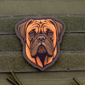Pode incluir: Um patch com uma ilustração detalhada do rosto de um cão, principalmente em tons de castanho e laranja, com um contorno escuro. O patch é fixado a um fundo de tecido verde com fechos de velcro.