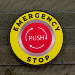 Könnte beinhalten: Ein runder Aufnäher mit den Worten "EMERGENCY STOP" in Schwarz auf gelbem Grund. In der Mitte befindet sich ein roter Kreis mit "PUSH" und Pfeilen. Der Aufnäher ist auf einem grünen Stoff befestigt.