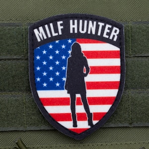 May include: A black and white patch with the text "MILF HUNTER" above a silhouette of a woman. The background of the patch is the American flag. The patch is on a green fabric.