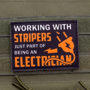 May include: A black and orange patch with the text "WORKING WITH STRIPERS JUST PART OF BEING AN ELECTRICIAN" and an orange electrical plug graphic. The patch is attached to a green fabric.