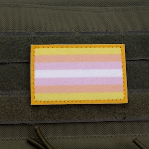 Op de afbeelding: Een rechthoekige patch met een genderqueer pride vlag ontwerp. De vlag heeft horizontale strepen in geel, wit, roze, oranje en geel. De patch is omrand met gele stiksels en staat tegen een donkergroene achtergrond.