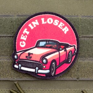 May include: A circular patch featuring a pink vintage convertible car on a bright pink background. The text GET IN LOSER is printed in white capital letters along the top curve of the circle. The patch has a black border and is attached to olive fabric.