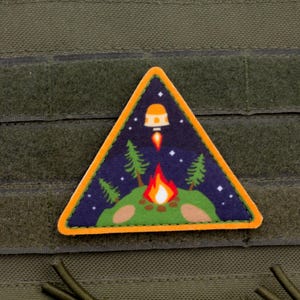 May include: A triangular patch depicting a night scene with a campfire, trees, and a rocket ship. The patch has an orange border and is set against a green background with velcro.