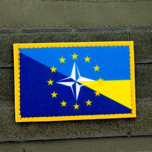 May include: A blue and yellow patch with a circle of stars and a white star in the centre. The patch is sewn onto a green fabric background.