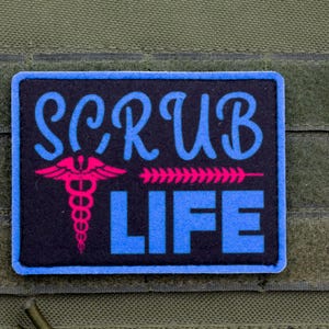 Può includere: Una toppa rettangolare nera con bordo blu che presenta il testo SCRUB LIFE in lettere blu. Un simbolo del caduceo rosa brillante è posizionato a sinistra della parola LIFE, e una fila orizzontale di frecce rosa punta verso destra.