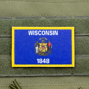 May include: A rectangular patch displaying the Wisconsin state flag, with a blue background and a yellow border. The patch shows the state seal, the word "WISCONSIN" and the year "1848" in white text. The patch is attached to a green fabric.