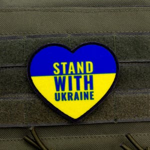 May include: A heart-shaped patch with the Ukrainian flag colors, blue and yellow, and the words "STAND WITH UKRAINE" in yellow. The patch is affixed to a green fabric background.