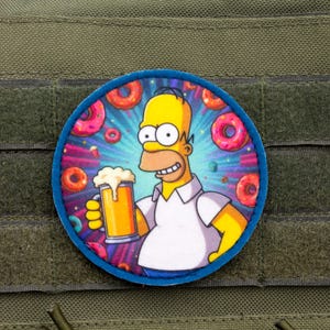 Puede incluir: Un parche redondo con Homer Simpson sosteniendo una jarra de cerveza, rodeado de rosquillas. El personaje de dibujos animados está sonriendo. El parche tiene un borde azul y está sobre un fondo verde.