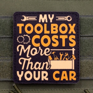 Può includere: Una toppa rettangolare nera con testo arancione e crema che recita MY TOOLBOX COSTS MORE THAN YOUR CAR. Il design include illustrazioni grafiche di chiavi inglesi, ingranaggi e una cassetta degli attrezzi piena di vari utensili manuali.