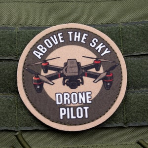 Può includere: Un cerotto circolare marrone e nero con il testo "ABOVE THE SKY DRONE PILOT" e un drone nero con accenti rossi al centro.