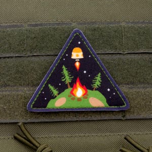 May include: A triangular patch depicting a night scene with a campfire, trees, and a rocket ship launching into space. The patch has a dark blue background with white stars and a green border. The patch is attached to a green fabric.