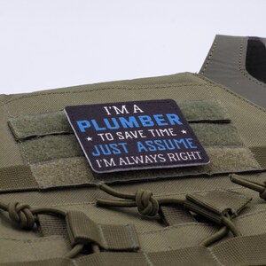 K&ouml;nnte beinhalten: Olivgr&uuml;ne taktische Weste mit einem rechteckigen Aufn&auml;her mit der Aufschrift "I'M A PLUMBER TO SAVE TIME JUST ASSUME I'M ALWAYS RIGHT" in wei&szlig;er und blauer Schrift. Die Weste hat mehrere Riemen und einen Klettverschluss.