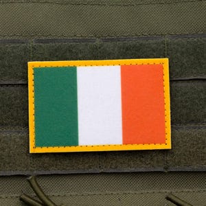 Può includere: Una toppa rettangolare con la bandiera irlandese, con strisce verticali verdi, bianche e arancioni, bordata di giallo. La toppa è attaccata a un tessuto verde oliva strutturato, probabilmente parte di un'attrezzatura tattica.