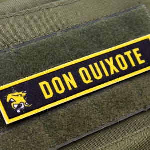 Puede incluir: Un parche de moral negro y amarillo con el texto "DON QUIXOTE" y una imagen estilizada de un caballo.