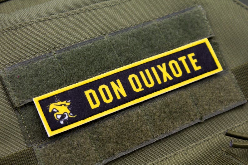 Puede incluir: Un parche de moral negro y amarillo con el texto "DON QUIXOTE" y una imagen estilizada de un caballo.