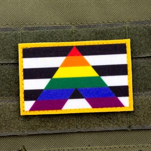 May include: A black and white striped patch with a rainbow triangle in the center. The patch has a yellow border and is attached to a green fabric background.