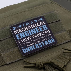 May include: A square patch with the text "I'M A MECHANICAL ENGINEER I SOLVE PROBLEMS YOU DON'T KNOW YOU HAVE IN WAYS YOU CAN'T UNDERSTAND" in white and blue on a black background. The patch is on a green fabric.