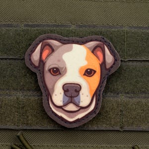 Op de afbeelding: Een patch met een gestileerd honden gezicht met een driekleurig ontwerp. Het gezicht van de hond is een mix van wit, grijs en oranje, met bruine ogen en een zwarte omtrek. De patch is bevestigd op een groene stoffen achtergrond.