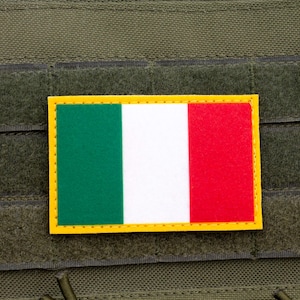 Può includere: Una toppa rettangolare con la bandiera italiana, con strisce verticali verdi, bianche e rosse e un bordo giallo. La toppa è attaccata a uno sfondo di tessuto verde, probabilmente un giubbotto tattico o attrezzature simili.