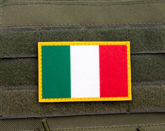 May include: A rectangular patch featuring the Italian flag, with green, white, and red vertical stripes, and a yellow border. The patch is affixed to a green fabric background, likely a tactical vest or similar gear.