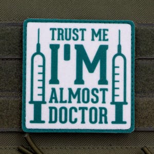Puede incluir: Un parche cuadrado con fondo blanco y borde verde azulado. El texto "TRUST ME I'M ALMOST DOCTOR" está impreso en letras de bloque verde azulado. A los lados izquierdo y derecho del texto, hay ilustraciones de líneas de jeringas médicas.