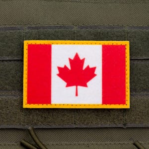 May include: A rectangular patch displaying the Canadian flag. The flag features a red maple leaf on a white background, with red vertical bars on either side. It has a yellow border and is attached to a green fabric.