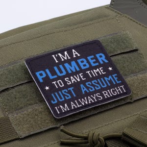 K&ouml;nnte beinhalten: Ein quadratischer Aufn&auml;her mit dem Text "I'M A PLUMBER TO SAVE TIME JUST ASSUME I'M ALWAYS RIGHT" in wei&szlig;er und blauer Schrift. Der Aufn&auml;her befindet sich auf einer gr&uuml;nen taktischen Weste.
