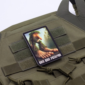 May include: Olive green tactical vest with a rectangular patch featuring a whimsical illustration of a forest creature with the text "BRR BRR PATAPIM". The vest has multiple straps and hook-and-loop fasteners.