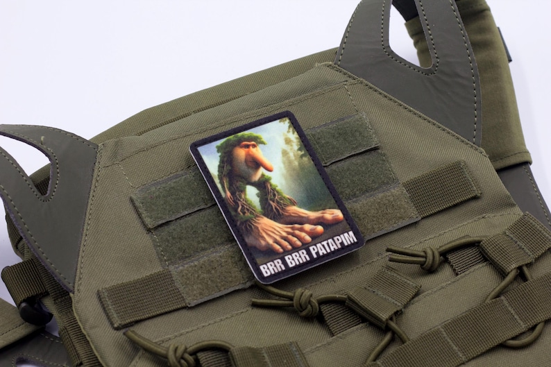 May include: Olive green tactical vest with a rectangular patch featuring a whimsical illustration of a forest creature with the text "BRR BRR PATAPIM". The vest has multiple straps and hook-and-loop fasteners.