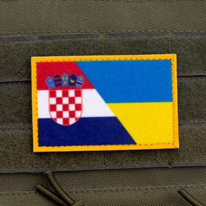 May include: A rectangular patch featuring a split design of the Croatian and Ukrainian flags, with a yellow border. The Croatian side has a red and white checkered emblem, while the Ukrainian side has blue and yellow stripes. The patch is attached to a green fabric.