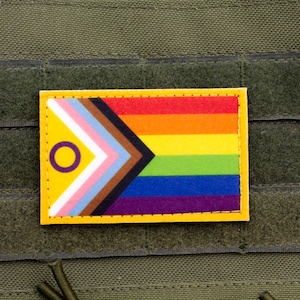 Peut inclure: Un patch jaune avec un motif de drapeau arc-en-ciel et un symbole de cercle avec un anneau violet autour. Le patch est sur un fond vert.